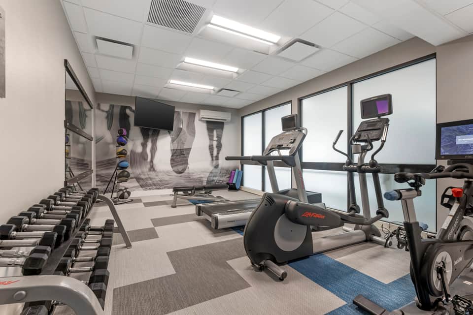 Modern fitness center with cardio equipment, treadmill, stationary bikes, dumbbells, and wall-mounted TV