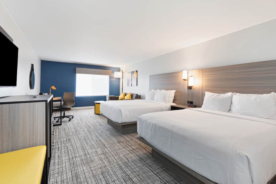 Modern hotel room with two queen beds, blue accent wall, work desk, and bright windows