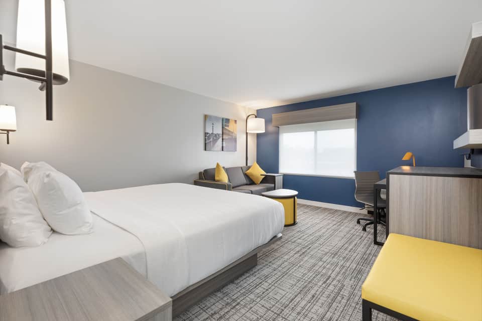 Spacious hotel room featuring queen bed, blue wall, yellow chair, and seating area
