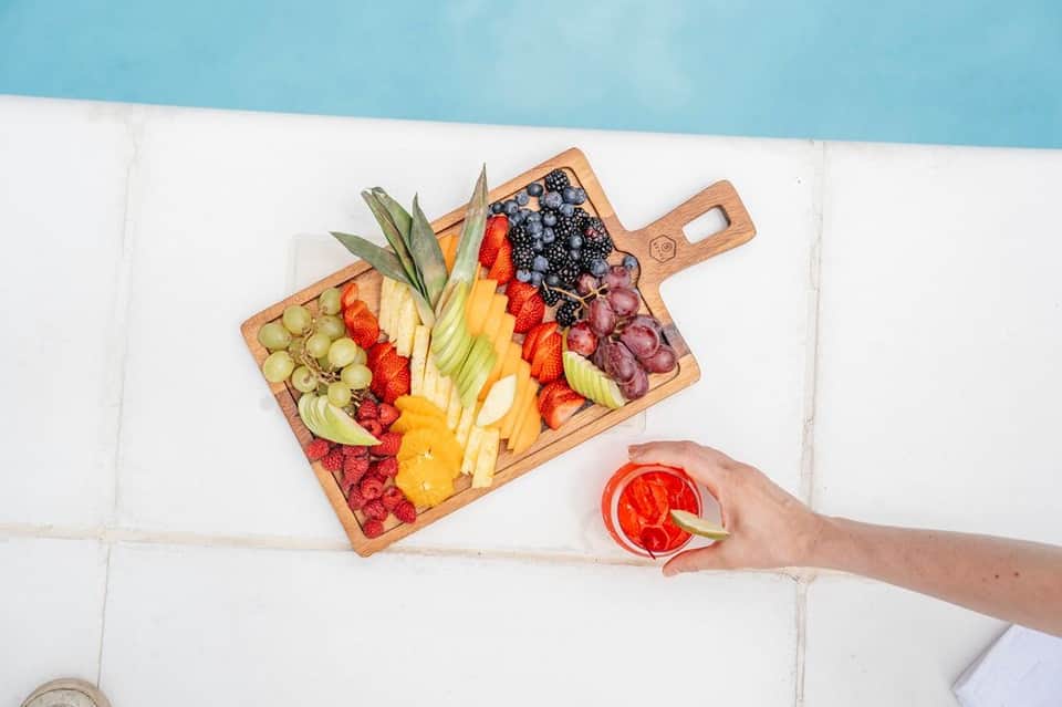 Wooden charcuterie board with fresh fruits, berries, and meats; hand holding grapefruit cocktail by pool