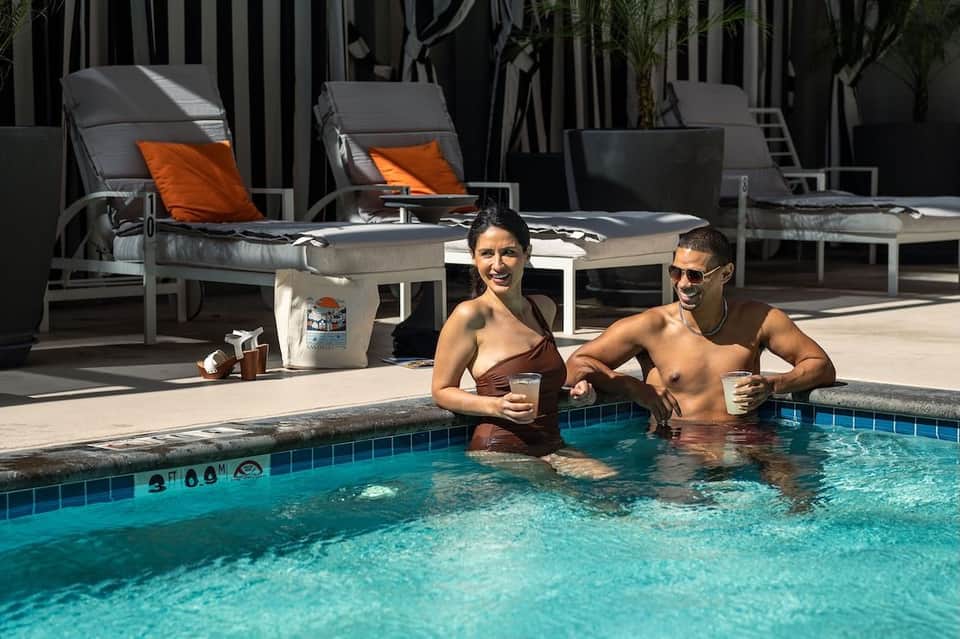 Two guests enjoying drinks in resort pool with loungers and striped umbrellas poolside