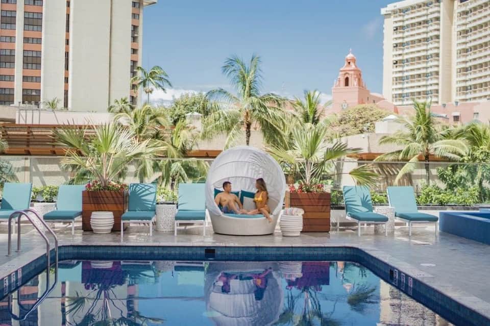 Resort pool deck with lounge chairs, white cabana, palm trees, and pink church dome visible in background cityscape