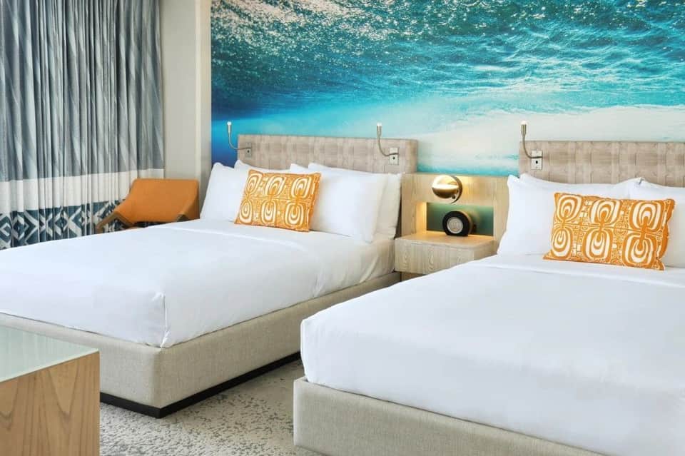 Modern hotel room with two queen beds, ocean beach mural headboard, white bedding, and gold accent pillows
