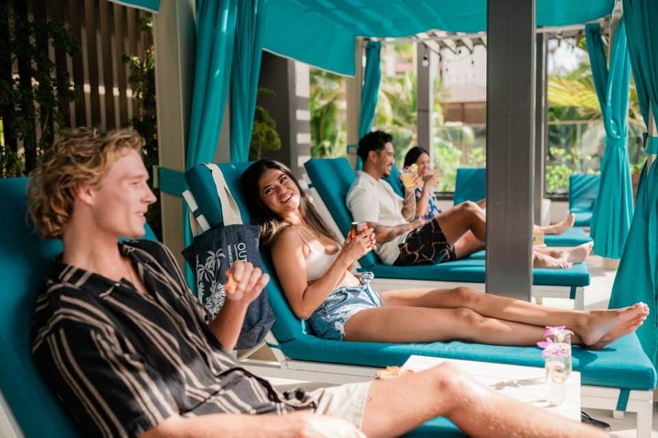 Guests relaxing on turquoise lounge chairs under teal canopy with curtains in shaded poolside area