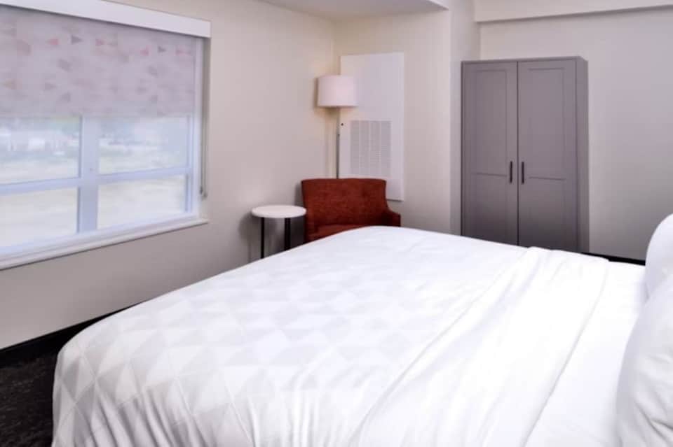 Modern hotel bedroom with queen bed, window, chair, lamp, and closet in neutral tones