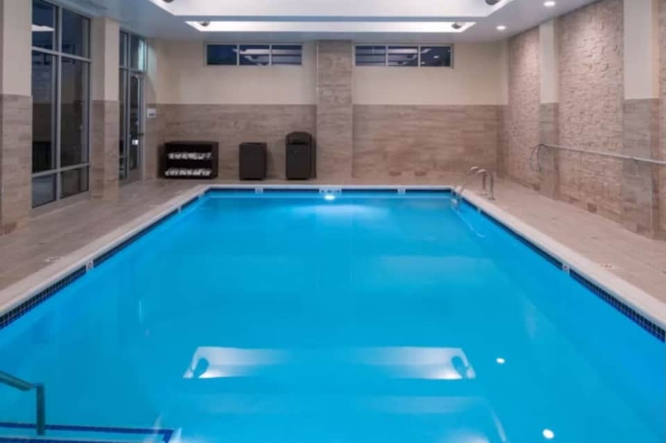 Indoor lap pool with brick walls, diving board, lounge chairs, and modern lighting fixtures