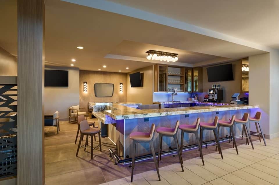 Modern hotel bar with illuminated counter, high seating, and multiple TV screens overhead