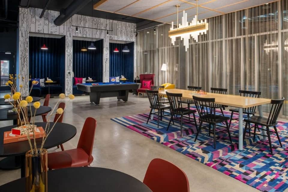 Hotel entertainment space with pool table, dining area, colorful geometric rug, and floor-to-ceiling curtains with neon accents
