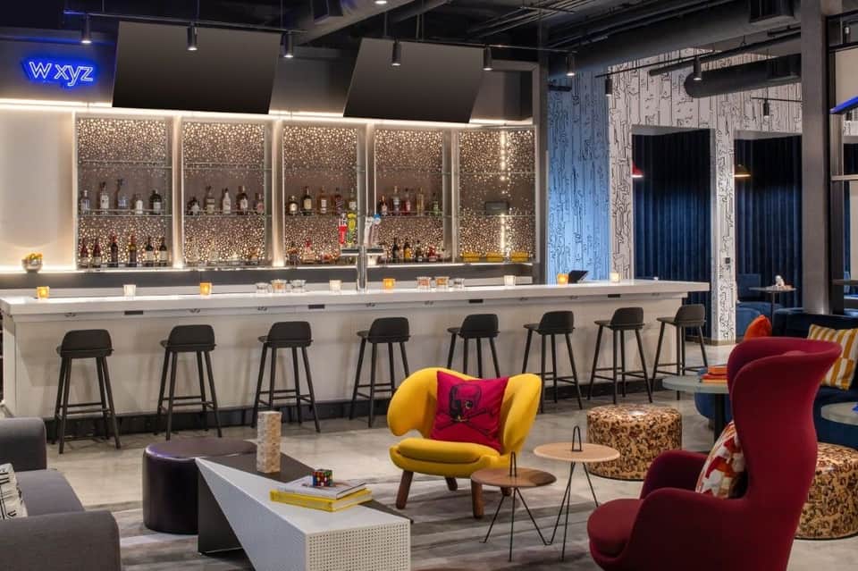 Modern hotel bar with illuminated bottle display, yellow and burgundy accent chairs, and contemporary lounge seating