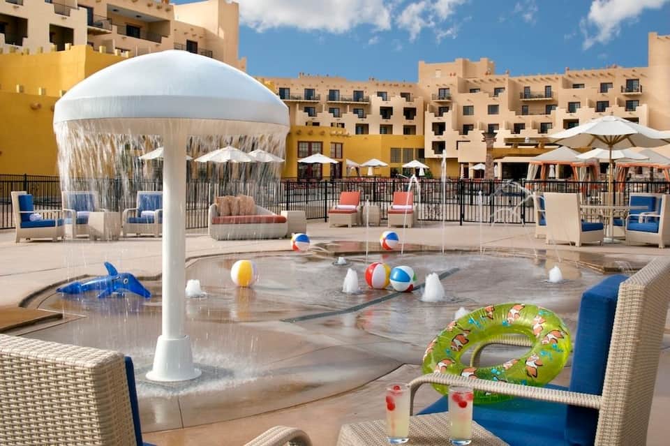 Kids' splash pad with white umbrella structure, colorful beach balls, and resort buildings in background