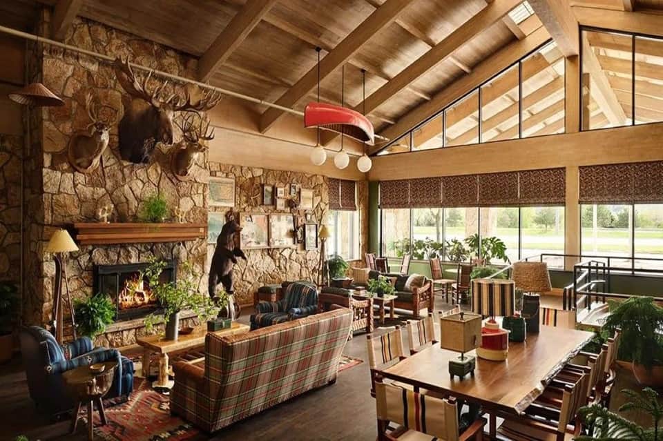 Rustic lodge interior with stone fireplace, wooden beams, plaid furniture, and expansive windows