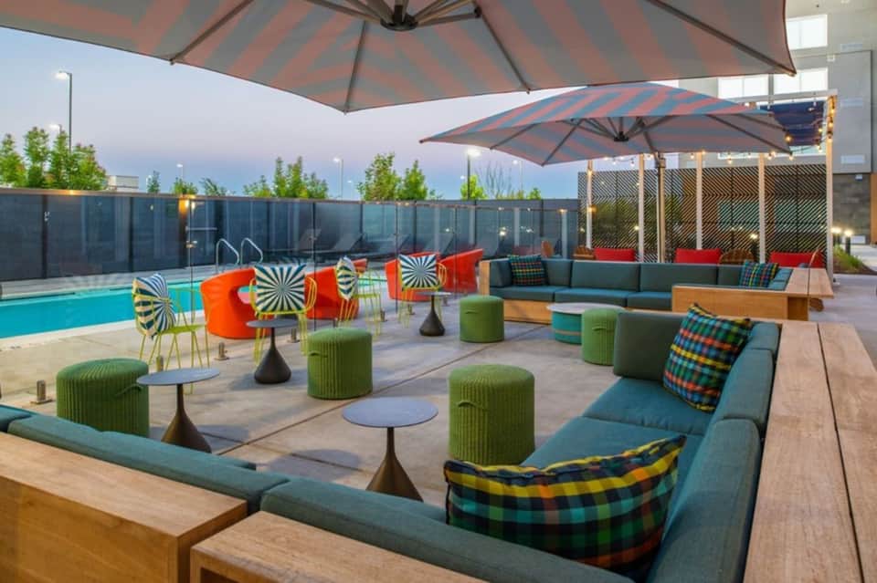 Pool deck lounge area at dusk with cabanas, teal sofas, green textured ottomans, striped umbrellas, and patterned throw pillows