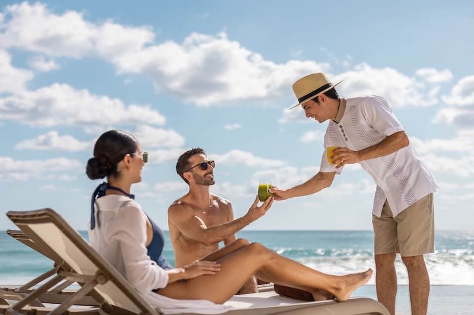 Server in white shirt and hat bringing drinks to couple relaxing on beach loungers