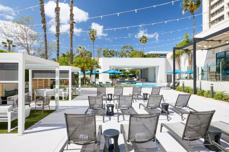 Outdoor resort deck with lounge chairs, palm trees, string lights, cabanas, and pool in background under blue sky
