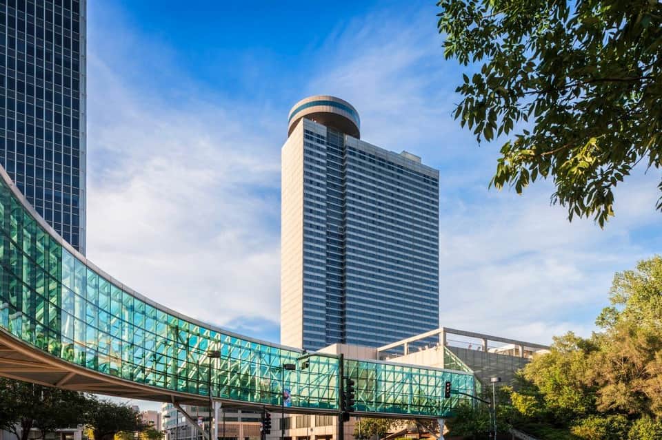 Modern cylindrical tower with glass pedestrian bridge and surrounding buildings under blue sky