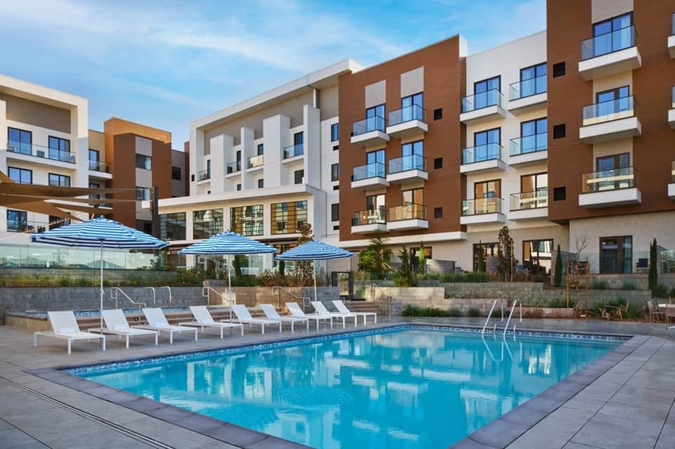 Resort swimming pool with blue umbrellas, loungers, and multi-story brick building complex