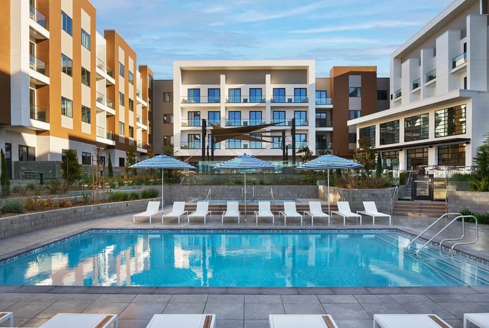 Ground-level pool with blue umbrellas and loungers surrounded by modern apartment buildings