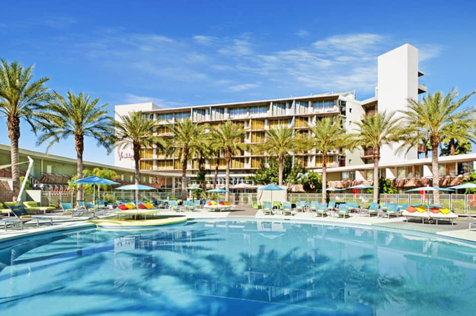 Resort pool with loungers and palm trees, modern white hotel building in background under blue sky