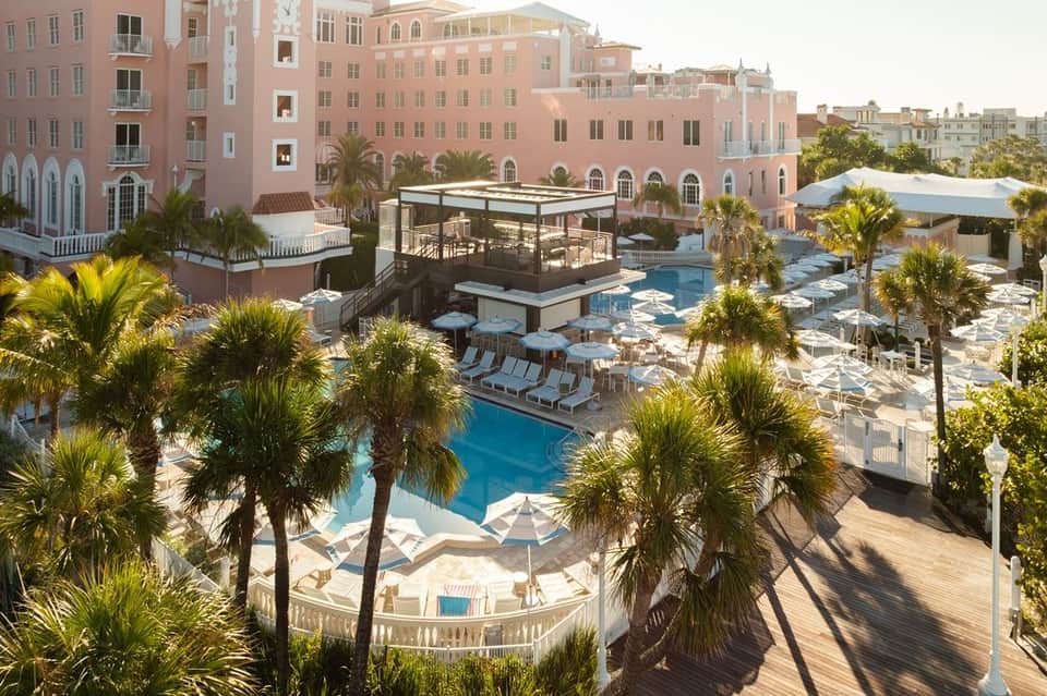 Aerial view of pink hotel with multiple pools, white umbrellas, and surrounding palm trees