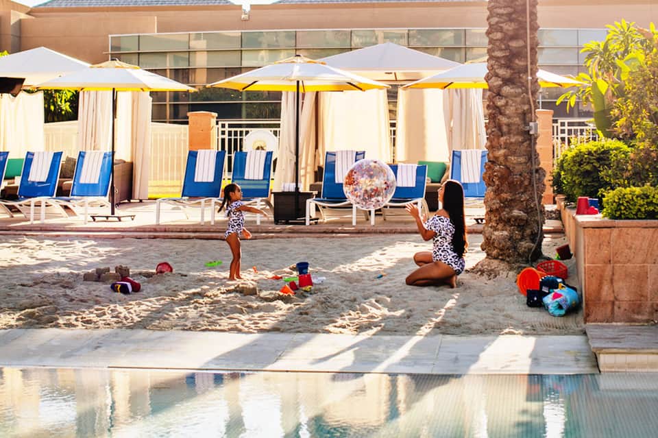 Mother and child playing with inflatable beach ball on sand near pool with lounge chairs and umbrellas