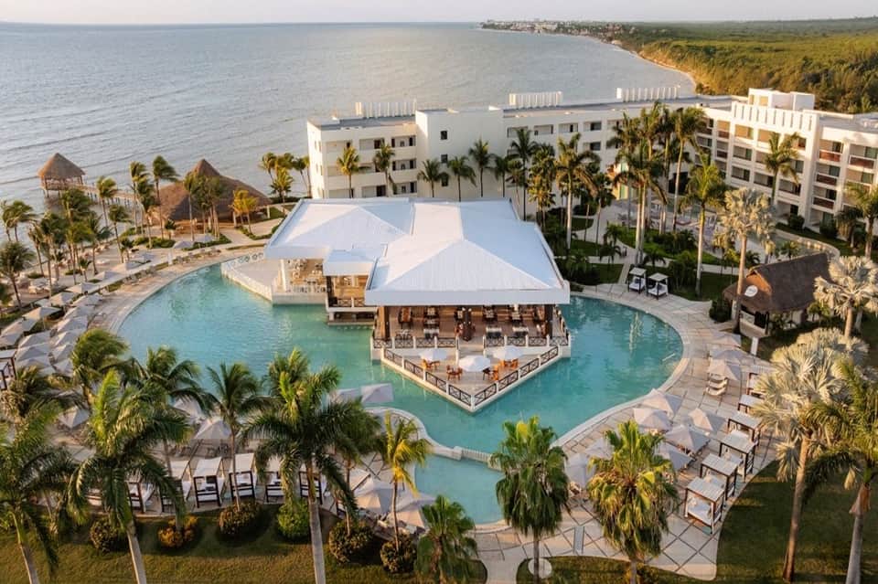 Aerial resort view with central lagoon pool, white roof structure, palm trees, and beachfront