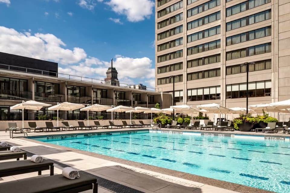 Outdoor lap pool with lounge chairs, white umbrellas, urban skyline backdrop, and clear blue sky