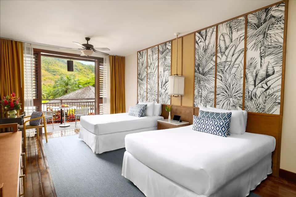 Spacious resort room with two queen beds, tropical headboard art, balcony, and jungle mountain views