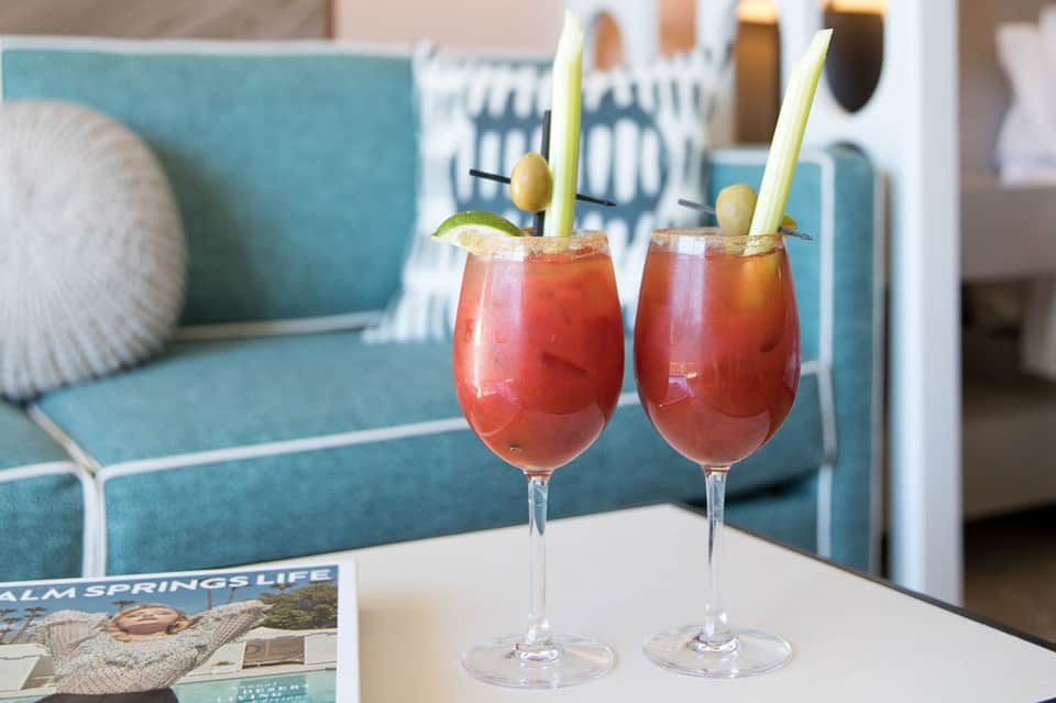 Two red cocktails with olives and green straws on white table with turquoise sofa in background