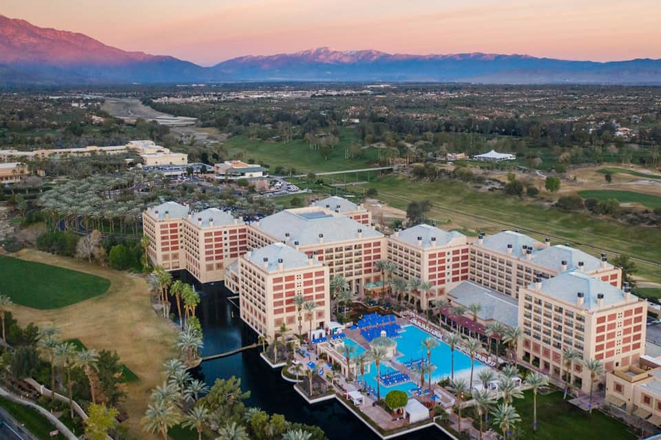 Aerial view of large pink resort with blue pool, palm trees, and snow-capped mountains in background at sunset