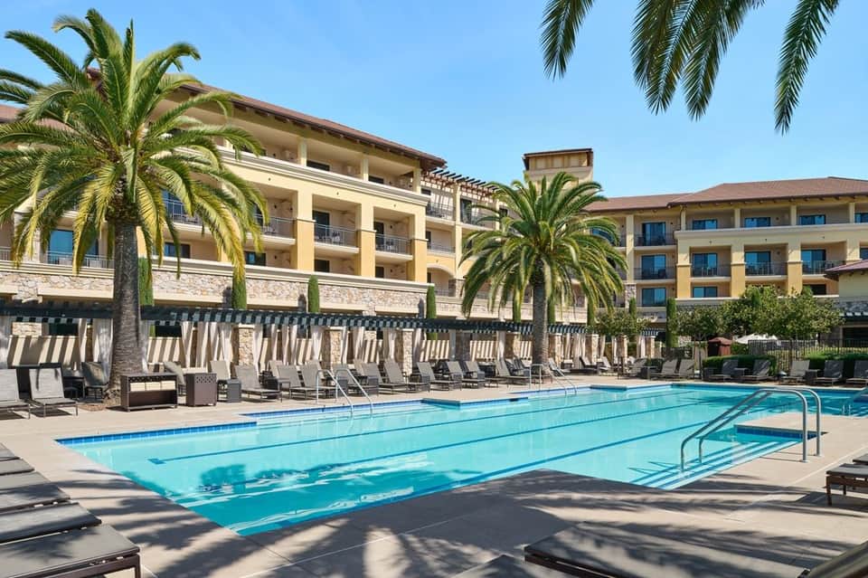 Resort swimming pool with multiple lounge chairs, palm trees, and multi-story hotel buildings in the background