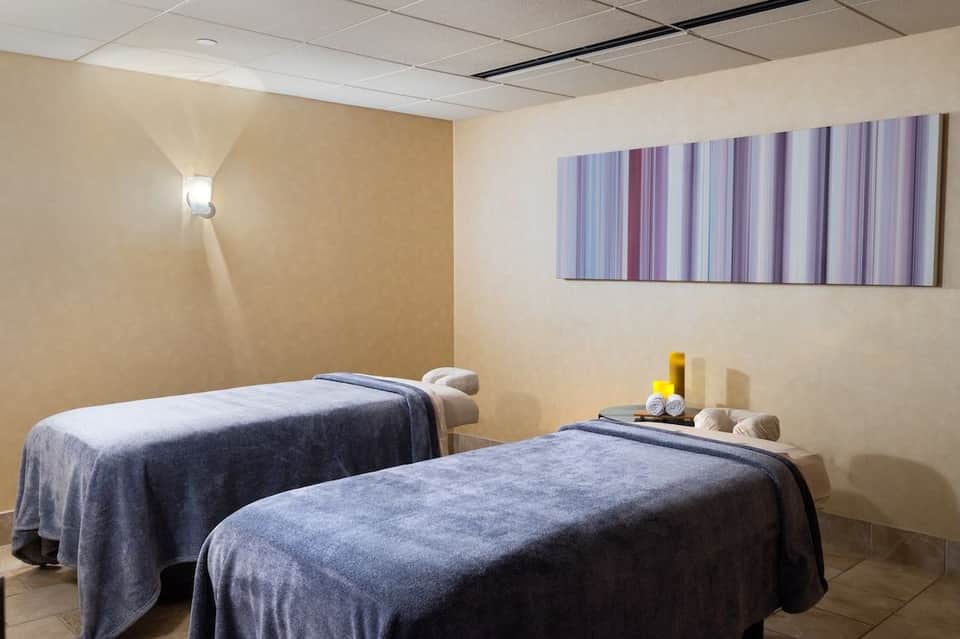 Hotel spa massage room with two treatment beds, neutral wall colors, and therapeutic supplies on shelf