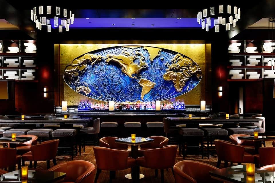 Upscale hotel bar lounge with artistic world map backdrop, modern chandelier, leather seating, and ambient lighting