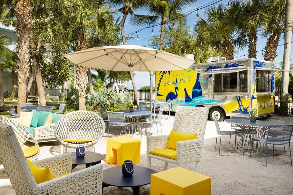 Outdoor lounge with yellow food truck, cream umbrella, woven seating, and palm trees