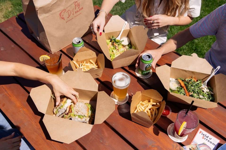Outdoor picnic table with burgers, fries, salads, and beers in cardboard containers on grass.