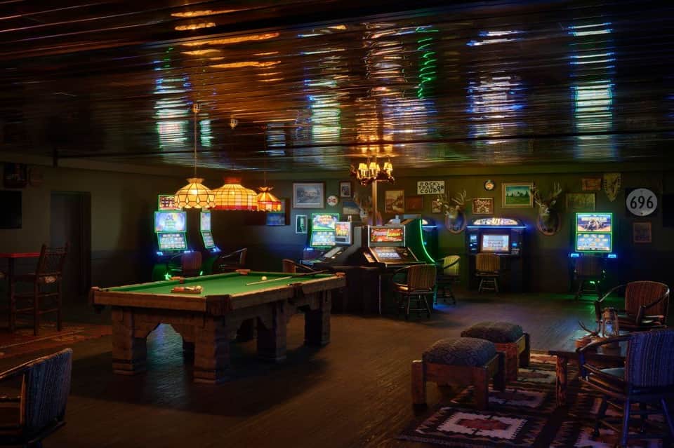 Dimly lit game room with pool table, slot machines, neon signs, and wooden furnishings.