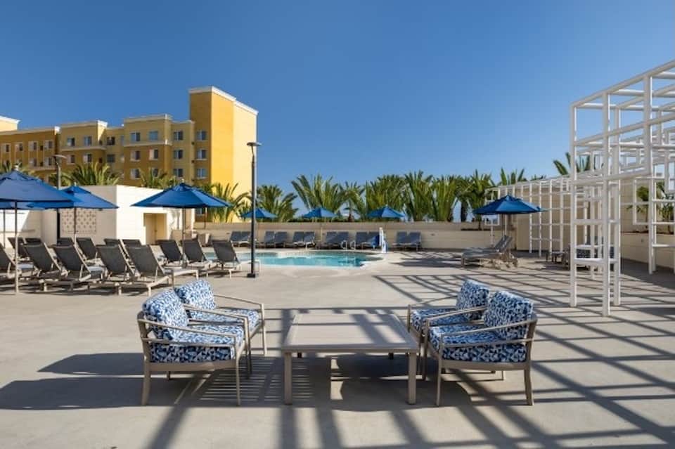 Resort pool deck with blue umbrellas, lounge chairs, white pergola, and palm trees