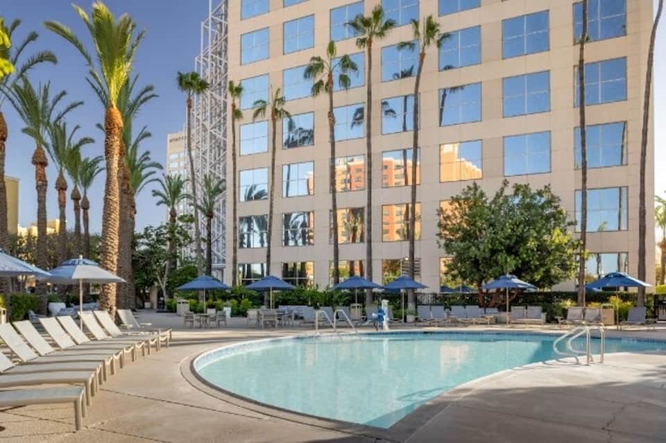 Resort pool with blue umbrellas and lounge chairs, palm trees, and modern high-rise hotel building in background