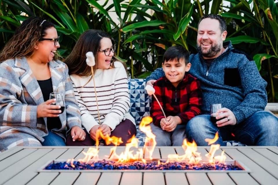 Family of four smiling around outdoor fire pit with blue umbrellas and green plants