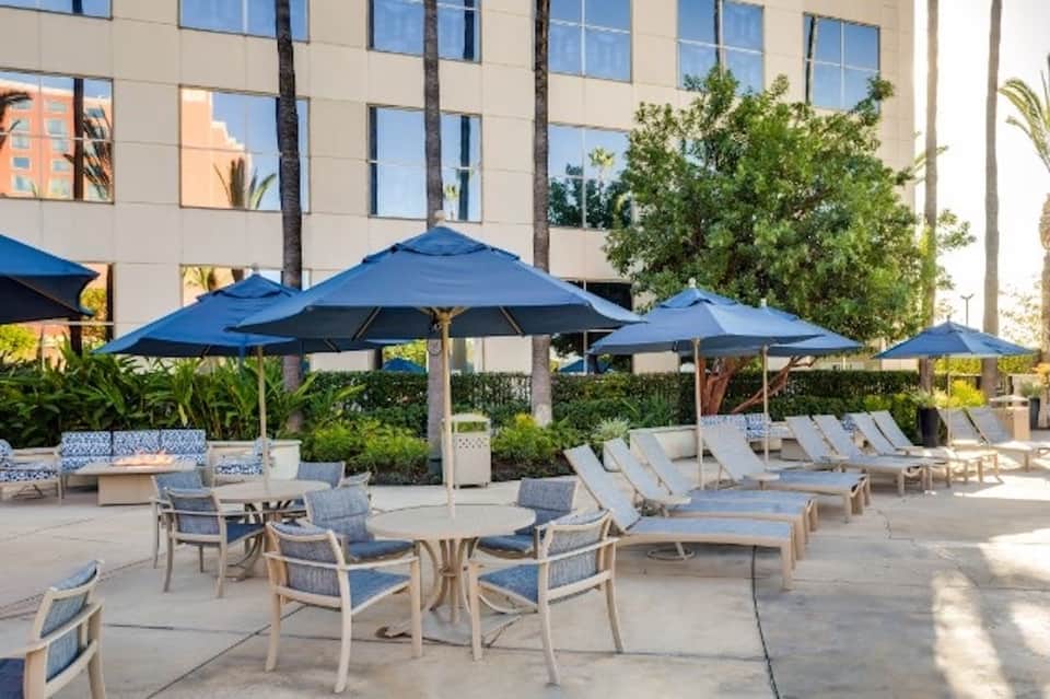 Poolside lounge area with blue umbrellas, modern hotel tower, palm trees, and seating arrangements
