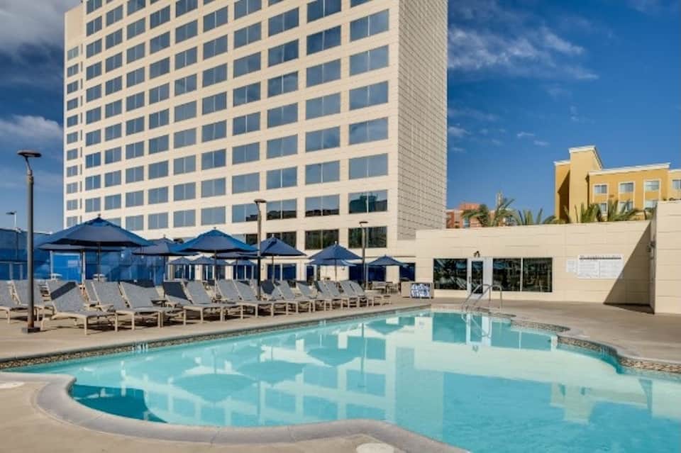 Resort pool with blue umbrellas, lounge chairs, modern high-rise hotel, and landscaped grounds