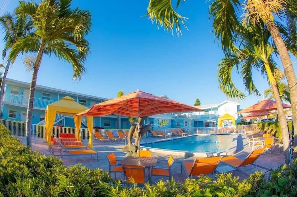 Resort pool with orange and yellow umbrellas, palm trees, turquoise water, and blue buildings.
