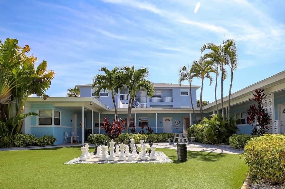 Multi-building pastel resort complex with giant chess set on lawn, palm trees, and clear sky.