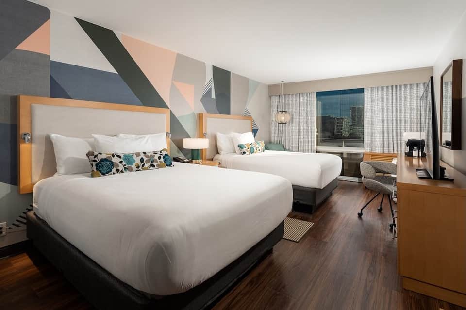 Modern hotel bedroom with two queen beds, geometric wall art, desk, and city view windows