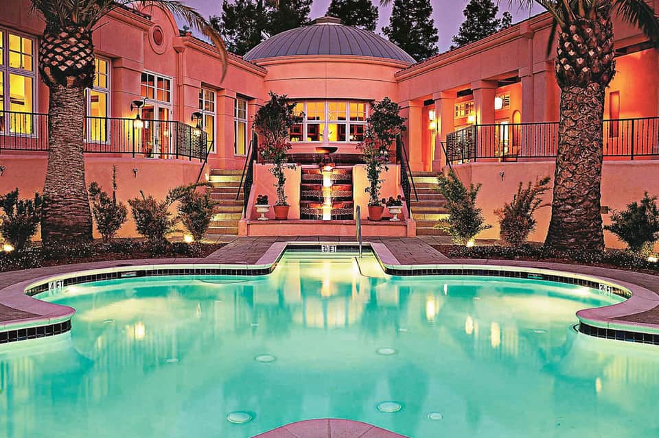 Illuminated courtyard with large turquoise pool, coral-pink building, palm trees, and evening sky