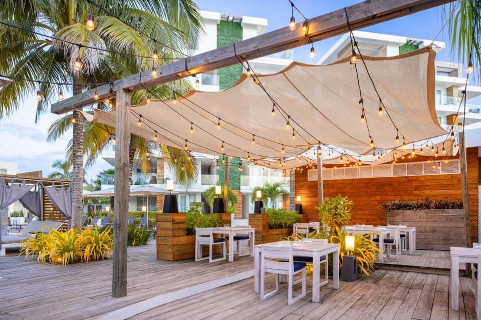 Outdoor dining area with wooden deck, shade sails, hanging lights, palm trees, and modern architecture