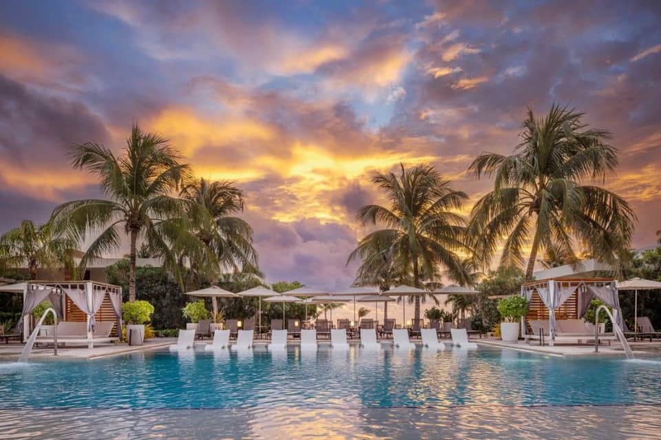 Luxury poolside at sunset with cabanas, palm trees, dramatic sky, and evening lighting