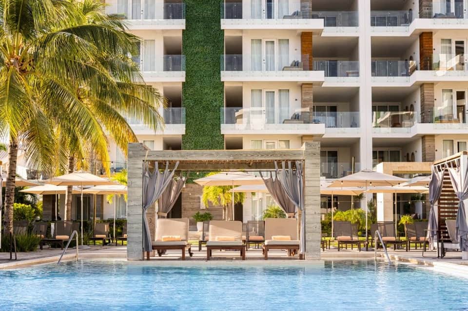 Modern resort pool area with white umbrellas, wooden pergolas, multi-story building, and palm trees