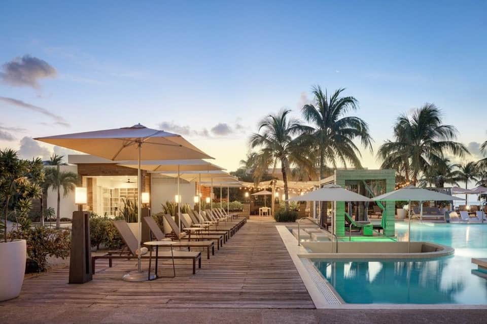 Resort pool deck at dusk with lounge chairs, large umbrellas, modern architecture, and palm trees