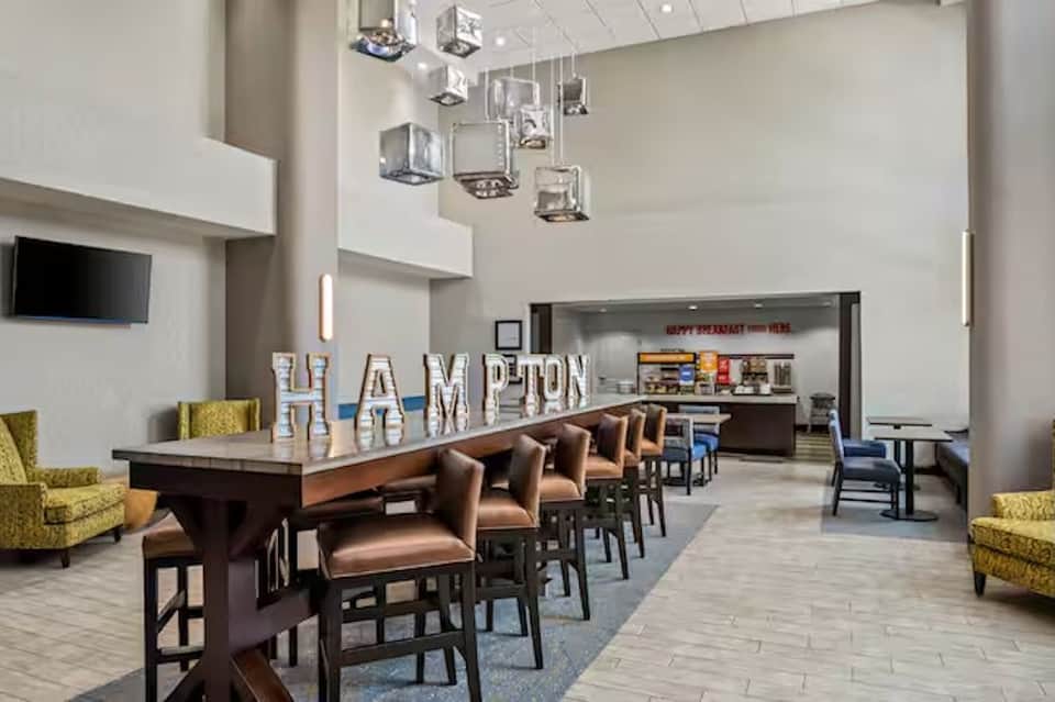Hotel lobby and breakfast area with high ceilings, HAMPTON sign, bar seating, and modern décor