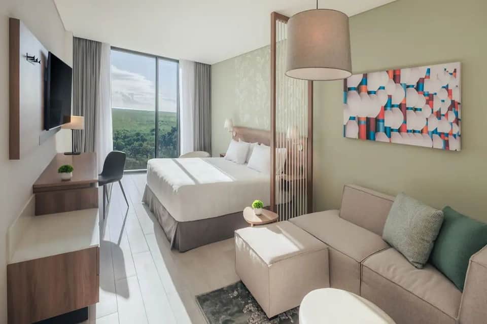 Hotel room with bed, sofa, work desk, TV, and large window overlooking green landscape